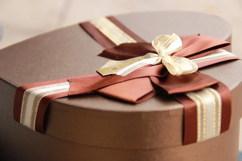 Nell'immagine un pacco regalo a forma di cuore color marrone con un nastro con fiocco a righe bianco, bordeaux e oro.