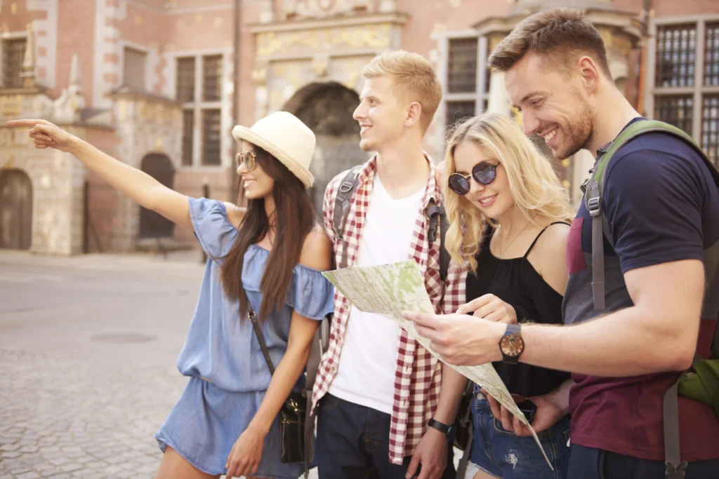 In the picture, a group of four young people is in a city and are visiting it. A girl points to a distant point to the boy next to her, while another boy and a girl look at a map.